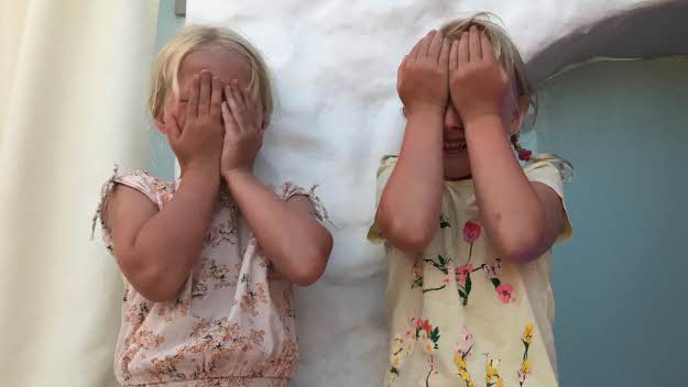 Två flickor sitter med händerna för ansiktet mot en vit mur.