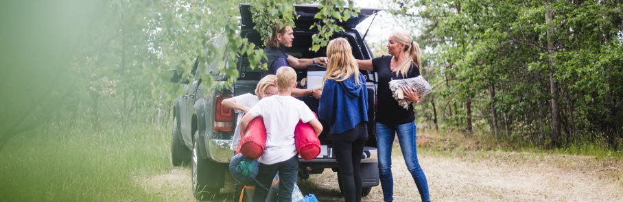 Familj vid bilen packar ut för camping i skogen.