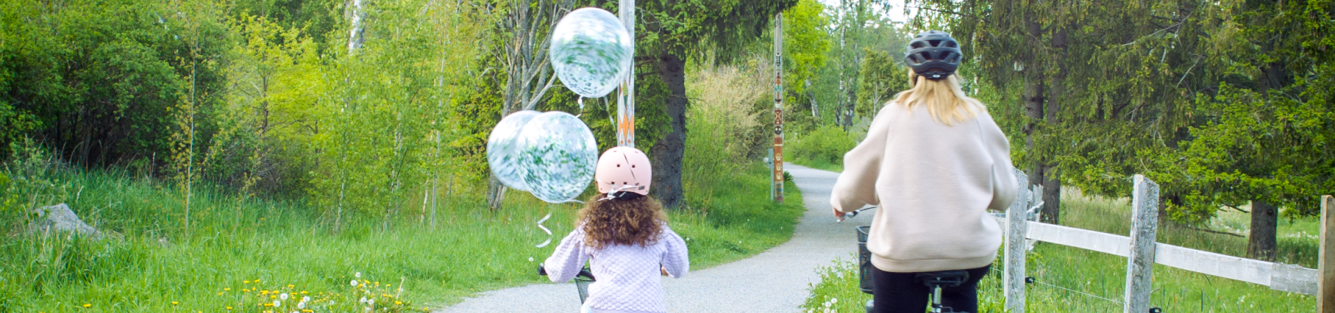 Cyklande mamma och dotter bakifrån på grusväg med grönska runtomkring och ballonger fladdrande från flickans cykel.
