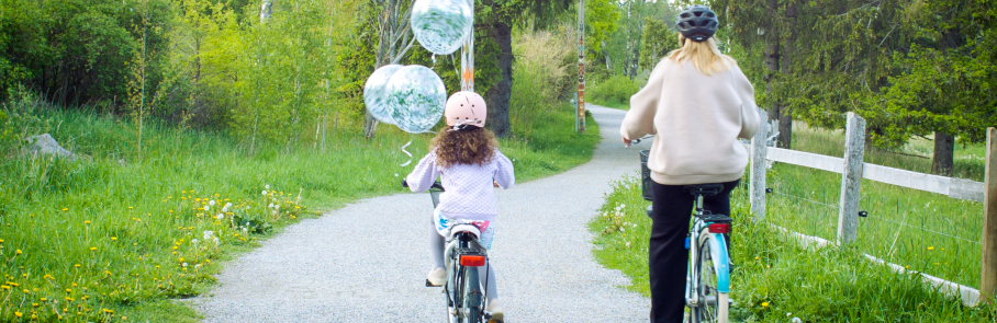 Cyklande mamma och dotter bakifrån på grusväg med grönska runtomkring och ballonger fladdrande från flickans cykel.